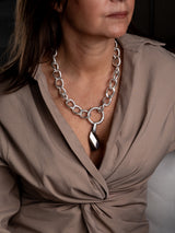 A close-up of a woman wearing a chunky silver chain necklace with a large central ring and a faceted teardrop pendant, paired with a taupe wrap blouse.