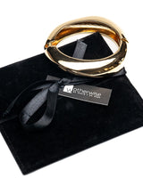 A chunky gold-toned metal ring bangle with a twisted, interlocking design, displayed on a black velvet presentation pouch with a black ribbon and a 'Wotherwise' brand tag.