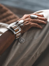 Chunky silver cuff bracelet with brown snake leather, five silver charms, worn on a wrist resting on light fabric.