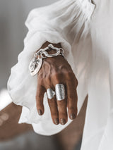Chunky silver-plated shackle bracelet with large coin pendants, worn on a hand with two silver rings, styled with a white flowy sleeve.