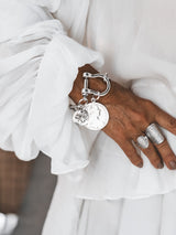 Chunky silver-plated shackle bracelet with large coin pendants, showcased on a wrist, paired with silver rings and a white blouse.