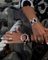 Black leather cuff bracelet featuring shiny oval metal elements, worn on a wrist beside other silver ring accessories.