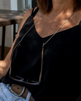 Thin, minimal silver chain for glasses worn around the neck, holding stylish sunglasses; model dressed in a black top.