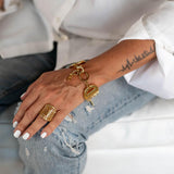 Chunky gold-plated stainless steel adjustable ring worn on a hand with a tattoo, paired with a golden bracelet and distressed jeans.
