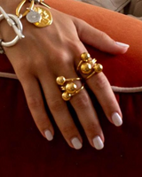 Adjustable stainless steel ring with large gold spheres, worn on two fingers alongside other jewelry, set against a soft, orange background.