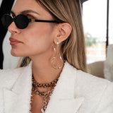 Large asymmetrical hammered stud earrings in shiny rose gold, worn by a model with layered necklaces and black sunglasses.