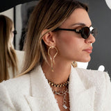 Large asymmetric dangle earrings with hammered irregular lines in rose gold, worn by a model with long hair and sunglasses.