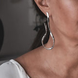 Large silver asymmetric earrings with exaggerated irregular lines, worn dangling from the model's ear.