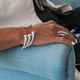 Silver-plated open bangle bracelet with a sleek, smooth design, worn alongside other bracelets on a hand resting on textured fabric.