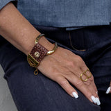 Chunky brown leather bracelet with a gold-plated cuff and decorative clasp, worn on a wrist with gold rings.