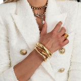 Three chunky gold metal cuff bangles worn stacked on a wrist, styled against a textured white blazer.