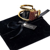 Unisex gold-plated bracelet featuring woven brown leather, displayed on a black suede pouch with a ribbon and logo tag.