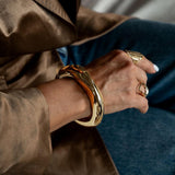 Chunky gold cuff bangle worn on the wrist, showcasing a high-polished surface, paired with additional rings on fingers.