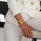 Chunky gold metal cuff bangles stacked on a wrist, showcasing a sleek design and polished finish, paired with a white blazer.