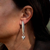 Asymmetric stainless steel earrings featuring a long pointed shape and a dangling heart, worn on the model's ear.