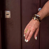 Brown leather strips form a chunky bracelet accented with a large gold metal plate, styled on a hand with a gold ring.