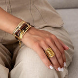 Chunky gold metal bracelet with brown leather strips worn on a model's wrist, paired with a gold statement ring.