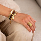 Chunky gold bracelet with large metal plate and brown leather strips, worn on a wrist alongside a matching gold ring.