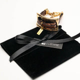 Chunky bracelet featuring a large gold metal plate and soft brown leather strips, presented with a suede gift pouch.