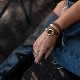 Brown leather wrap bracelet with honey gold metal links, stacked with other bracelets, worn on a hand with a gold ring.