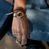 Honey gold metal and brown leather wrap bracelet with chunky links, layered with dark leather bands and gold rings.