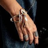 Brown suede leather bracelet featuring a silver horsebit buckle and tassel, worn on a wrist adorned with rings.