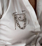 Silver safety pin brooch with chains and cross pendants attached, styled on a white button-up shirt.