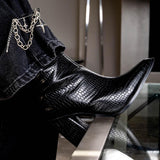 Silver safety pin brooch with chains and crosses attached, styled on black croc-embossed ankle boot, worn with dark jeans.