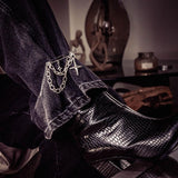 Silver safety pin brooch with chains and cross pendants attached to dark denim pants, paired with black textured boots.