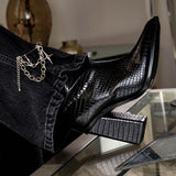 Silver safety pin brooch featuring chains and crosses, styled on black denim and paired with glossy black heeled boots.