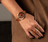 Chunky hammered gold hoop bracelet with natural leather strands, worn on a wrist with gold rings and dark nail polish.