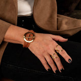 Chunky hammered gold hoop bracelet features natural leather strands, worn on a wrist with bold rings and dark nails.