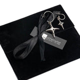 Silver hoop earring set featuring dangle cross and black enamel charms, displayed in a black suede pouch.
