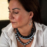 Silver hoop earrings with dangle cross and black enamel charms worn by a woman, layered with chunky beaded necklaces.