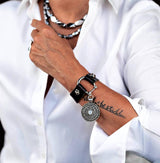 Person wearing a black leather bracelet with a silver charm on a white shirt background