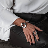Wide punk bangle bracelet with exaggerated irregular loop design, polished metal finish, worn on wrist with matching ring.