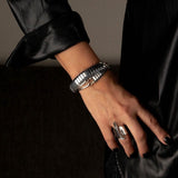 Hematite gemstone wrap bracelet with polished beads and silver-tone hardware, worn layered over a black leather sleeve.