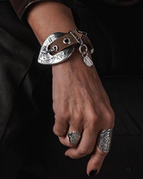 Khaki double-wrap leather bracelet with a large silver-plated western buckle, worn on a hand with chunky rings.