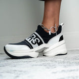 Chunky silver anklet with a T-clasp, worn with stylish black and white sneakers on a light textured surface.