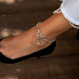 Stainless steel anklet with oval links, worn on the ankle, featuring a small charm and a minimalist design.