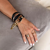 Double link metal bracelet featuring honey gold accents and a black leather wrap, worn stacked with rings on a hand.