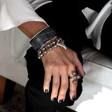 Wide brass cuff bracelet with dark brown animal print leather, worn stacked with silver beaded and ring accessories.