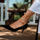 Silver snake chain anklet worn on a bare ankle, paired with a black pointed-toe heel and white frayed jeans.