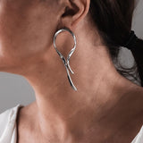 Asymmetric silver-tone dangle earrings featuring flowing, irregular wire shapes and hammered stud tops, worn on the ear.