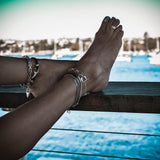 Silver and gun metal snake chain anklet showcased on bare feet, layered with other bracelets, against a blue water backdrop.