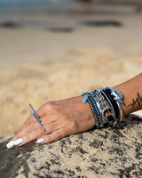 Geometric bar knuckle ring in silver worn on a hand with metallic bracelets, resting on a rock with a sandy beach background.