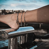 Three silver anklets stacked on an ankle, featuring delicate rhombus, bar link, and oval link designs in stainless steel.