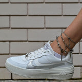 Three silver anklets layered together, featuring delicate rhombus, bar link, and oval link chains, worn with white sneakers.