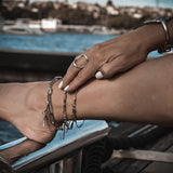 Three silver anklets stacked on an ankle, featuring rhombus, bar link, and oval link designs in stainless steel.