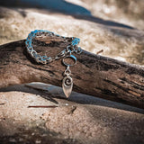 Stainless steel anklet featuring a solid square byzantine chain and an ornate pendant, resting on driftwood.